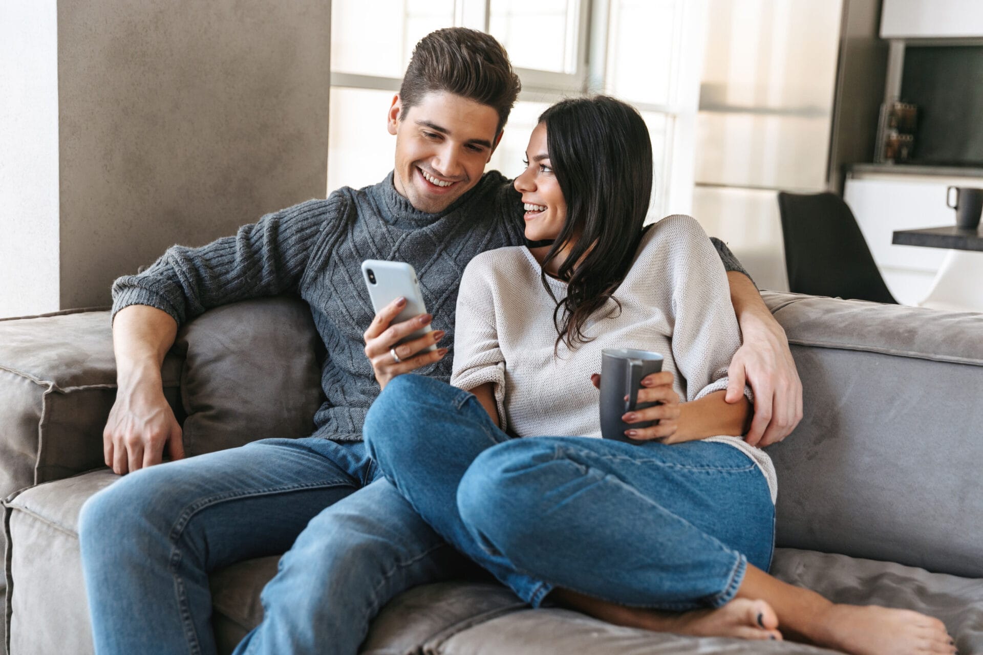 Happy young couple sitting on a couch at home, drinking tea, looking at mobile phone