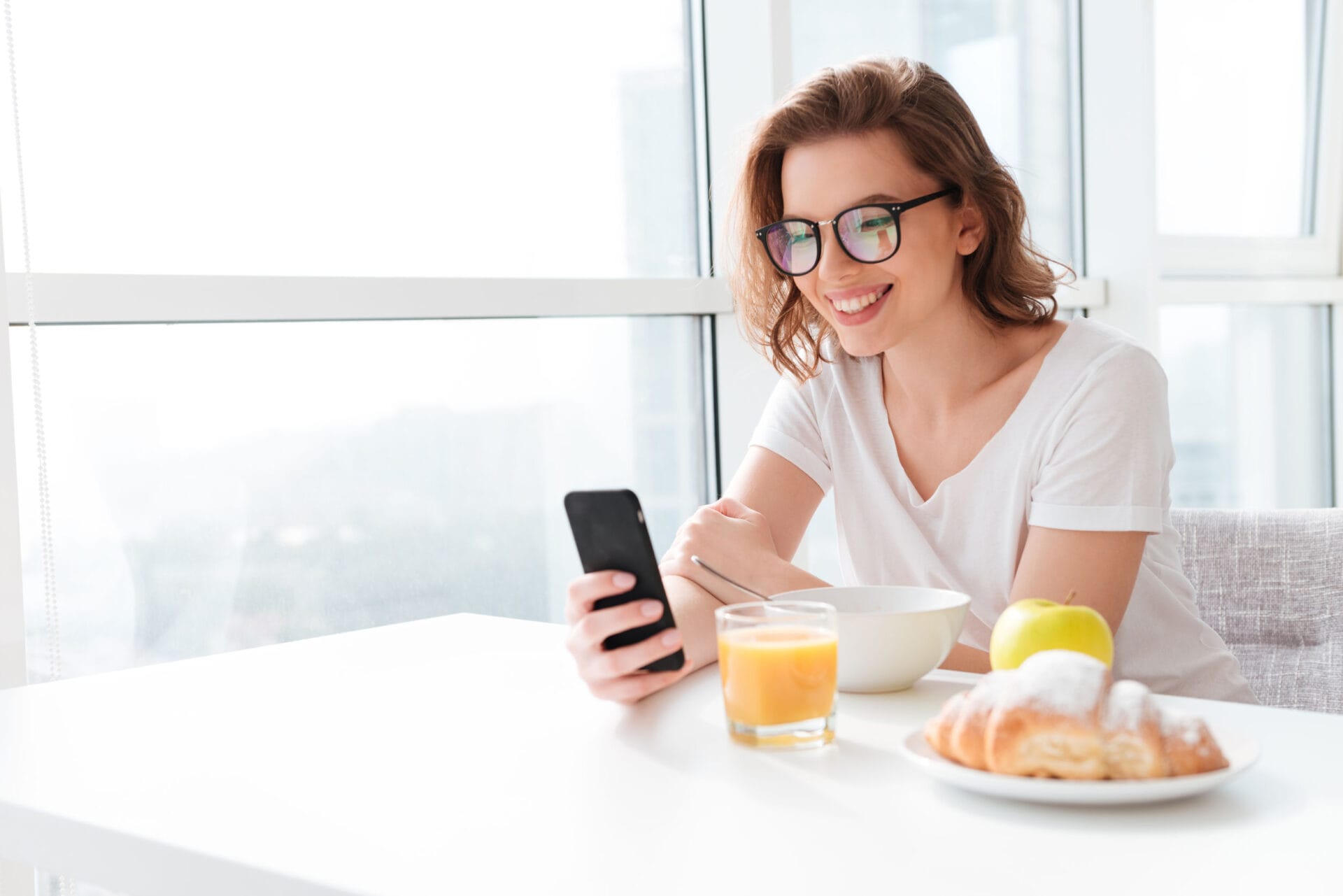 Photo of cheerful young amazing woman sitting indoors at the table with juice and croissant and corn flakes. Looking aside chatting by mobile phone.