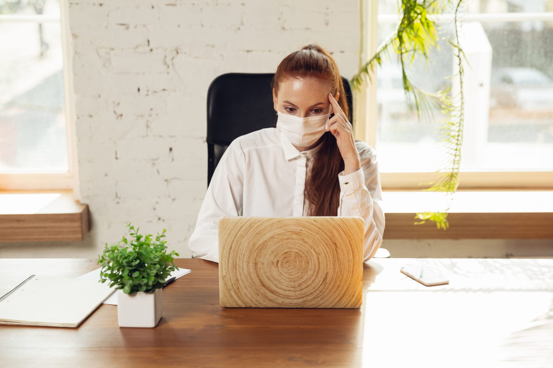 Getting info. Woman working in office alone during coronavirus or COVID-19 quarantine, wearing face mask. Young businesswoman, manager doing tasks with smartphone, laptop, tablet has online conference.